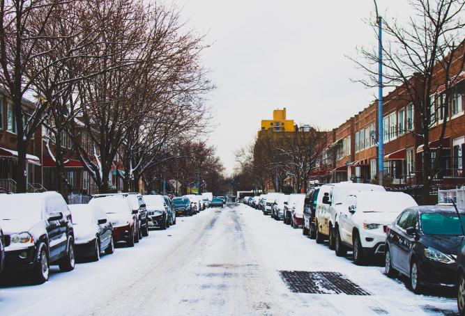 Snow Covered Road and Inline Parked Vehicles Between 2-storey Buildings Under White Sky